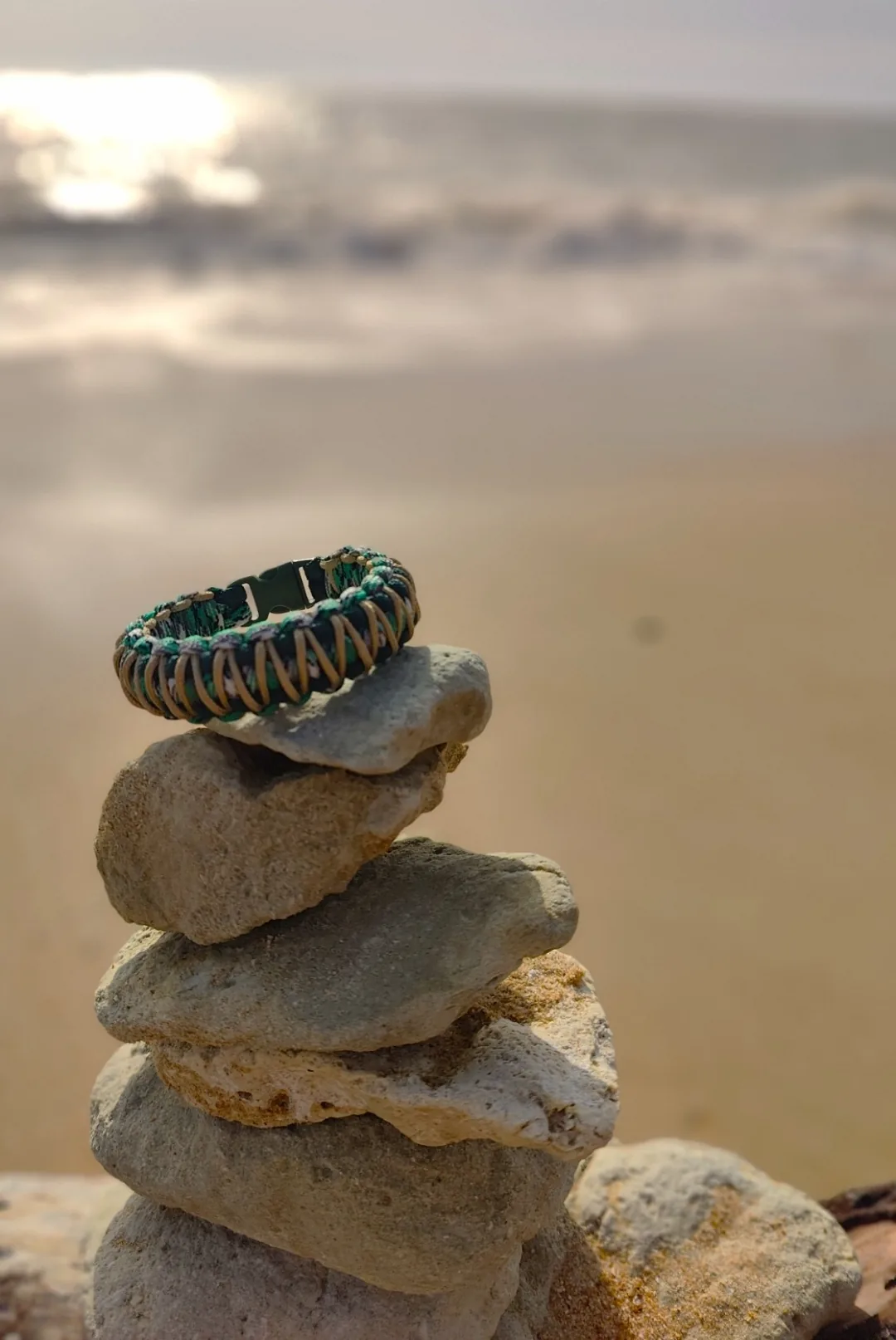 Bracelet vert, beige et brun en paracorde posé sur un cairn de pierres au bord de la mer