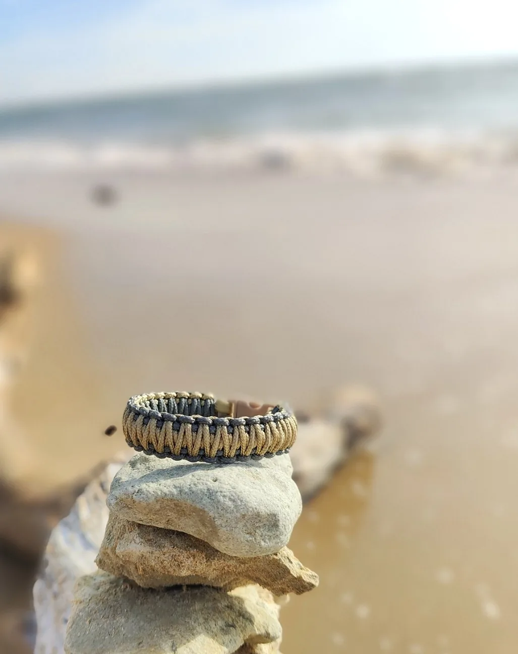 Bracelet beige et gris en paracorde posé sur des pierres avec la mer en arrière-plan