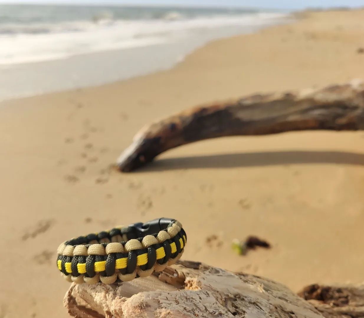 Bracelet jaune, beige et noir en paracorde présenté sur du bois flotté en bord de mer